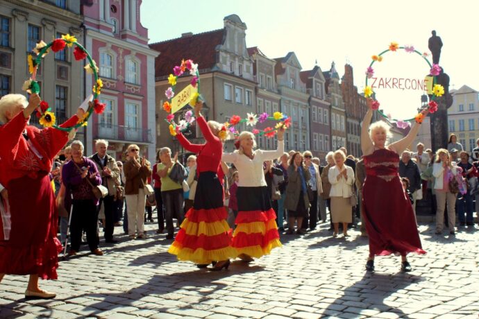 Seniorki z zespołu „Zakręcone babki”w kolorowych strojach tańczą na płycie Starego Rynku. 