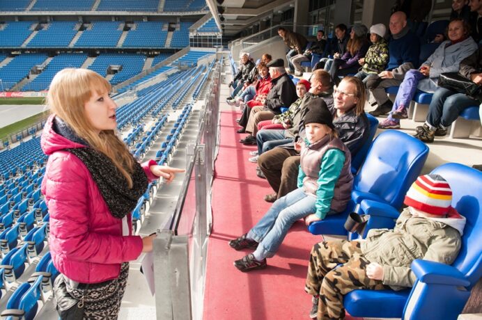 Na zdjęciu uczestnicy wycieczki po INEA Stadionie siedzą na trybunach. Prowadząca wycieczką stoi na przeciwko uczestników. 