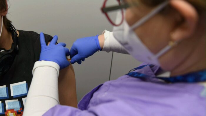 Zdjęcie pielęgniarki, widok z tyłu, w centrum fotografii jej dłonie w niebieskich rękawiczkach medycznych - wykonuje szczepienie w odkryte ramię pacjenta, fot. UMP