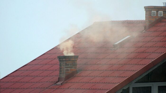 Grafika artykułu: zdjęcie czerwonego dachu z kominem, z komina wydobywa się żółtoszary dym.