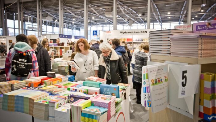 Grafika artykułu: zdjęcie Poznańskich Targów Książki na MTP - na pierwszym planie stosy książek, którym przyglądają się dwie kobiety, w tle szereg stoisk z książkami.