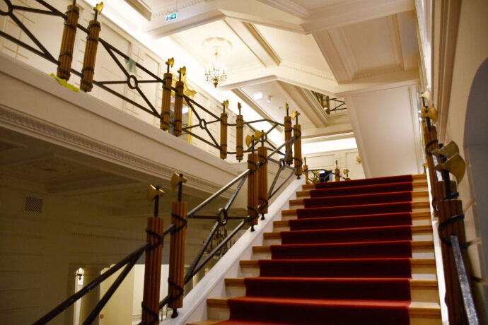 Grafika artykułu: Zdjęcie wnętrza historycznego gmachu biblioteki, na pierwszym planie schody pokryte czerwonym dywanem, w tle widać sufity z zabytkowymi żyrandolami. 