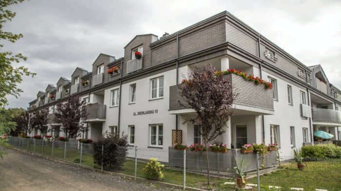Grafika artykułu: Na zdjęciu szaro-biały budynek przy ul. Drewlańskiej. Przed budynkiem trawnik z krzewami i drzewami, na balkonach budynku widać kolorowe kwiaty. 