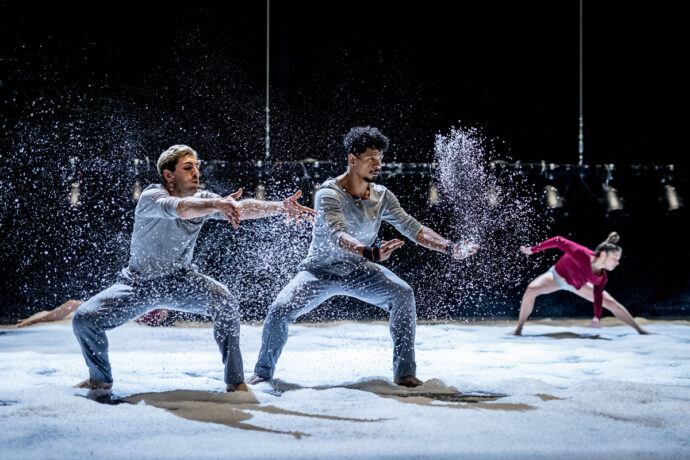 Grafika artykułu: Na zdjęciu na pierwszym planie dwoje tancerzy w szarych strojach, wokół nich biały pył, w tle tancerka w różowej bluzce, lekko pochylona. 