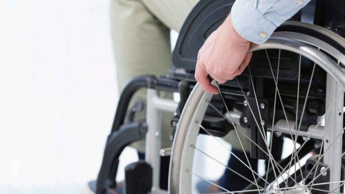 Grafika artykułu: zdjęcie osoby na wózku inwalidzkim - zbliżenie dłoni trzymającej koło wózka inwalidzkiego.