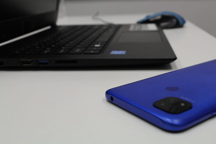 Grafika artykułu: Na zdjęciu, na pierwszym planie widać niebieski smartfon. W tle czarny laptop z niebieską myszką. Sprzęty leżą na białym stole. 