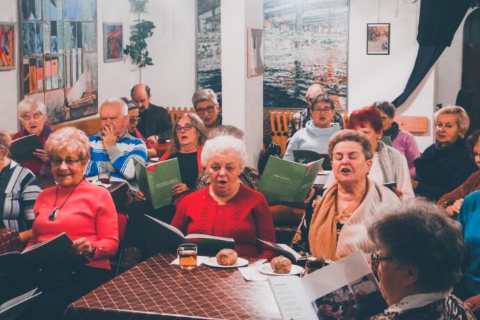 Grafika artykułu: Na zdjęciu widać śpiewające seniorki i seniorów, wszyscy siedzą przy stolikach, trzymają w rękach śpiewniki. 