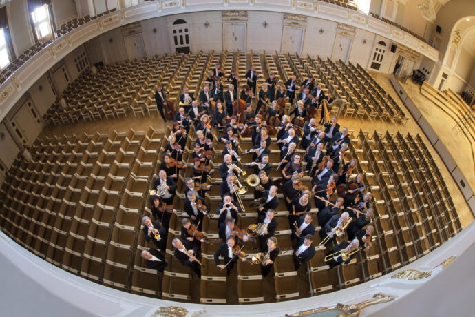Grafika artykułu: zdjęcie przedstawia orkiestrę symfoniczną Filharmonii Poznańskiej. Ujęcie zrobione jest od góry, z perspektywy osoby stojącej na balkonie. Członkowie orkiestry grają na instrumentach, ustawieni są pomiędzy siedzeniami dla publiczności.