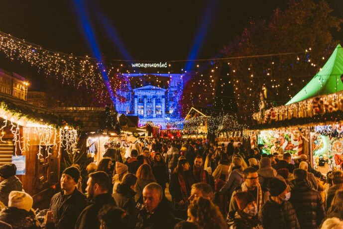 Grafika artykułu: zdjęcie przedstawia poznański Plac Wolności po zmroku. W oddali podświetlony budynek Arkadii z podświetlonym neonem "Wolność". Na placu rozstawione są drewniane, oświetlone jarmarczne stoiska/domki. Na środku widać tłum rozmawiających i przechadzających się ludzi. Cały plac rozświetlony jest lampkami bożonarodzeniowymi.