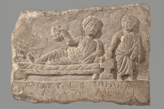 Grafika artykułu: stela grobowa, znajdują się na niej dwie postacie - jedna leży na łóżku, druga stoi obok, na dole starożytne napisy.