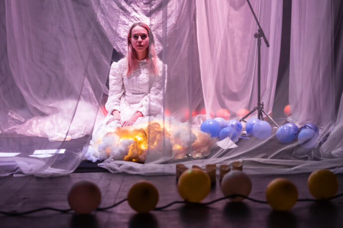 Grafika artykułu: Zdjęcie wykonane podczas spektaklu "Św. pępek świata", na pierwszym planie żółte balony, na drugim - aktorka w białej sukni, siedzi na scenie, za białą, przezroczystą zasłoną, wokół niej czerwone i niebieskie balony. 