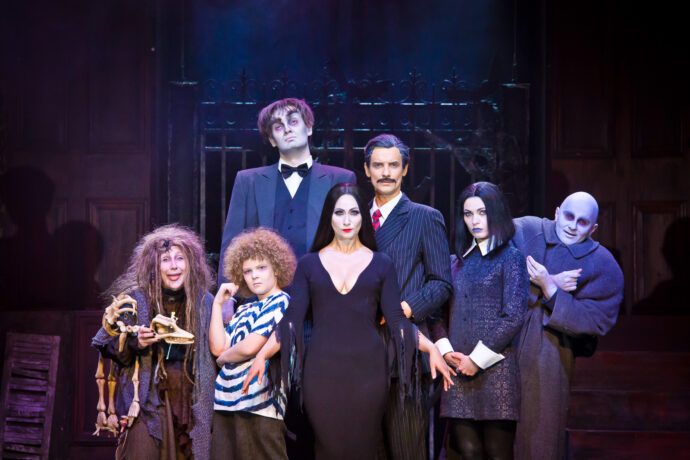 Grafika artykułu: Na scenie aktorzy grający główne postacie w spektaklu „Rodzina Addamsów”, patrzą na wprost, są oświetleni, ubrani na czarno, z wyjątkiem jednego chłopca, który ma koszulkę w niebiesko-białe paski. W tle żelazna brama w półmroku.