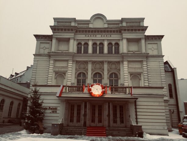 Grafika artykułu: Zdjęcie budynku Teatru Polskiego w Poznaniu, nad wejściem do teatru umieszczono powstańczy kotylion i flagi polskie, obok wejścia stoi choinka.