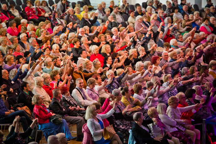 Grafika artykułu: Na zdjęciu seniorzy i seniorki siedzą na krzesłach, zdjęcie wykonane podczas koncertu w trakcie targów senioralnych. Niektórzy mają podniesione ręce, za które się trzymają.