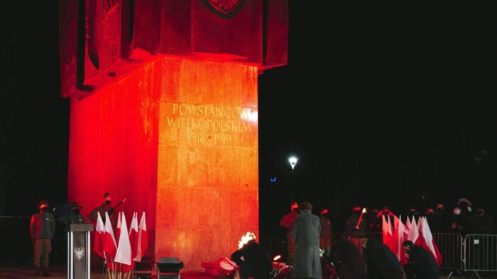 Grafika artykułu: w centrum pomnik z podświetlonym napisem "Powstańcom Wielkopolskim 1918-1919". Pięć osób składa przed pomnikiem wieńce. Zdjęcie wykonano po zmroku.