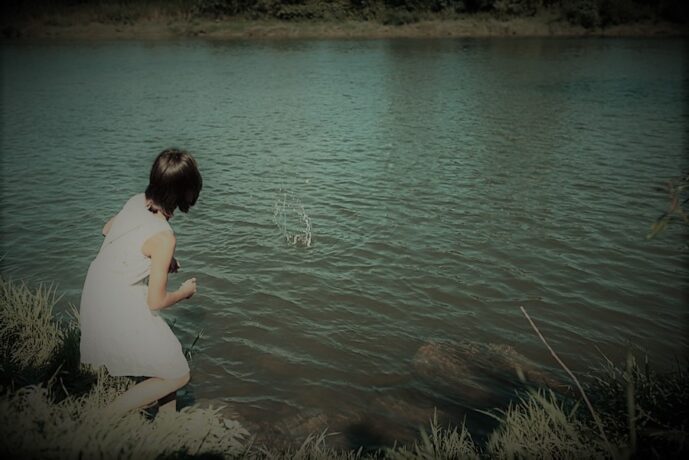 Grafika artykułu: Na zdjęciu dziewczynka stojąca nad brzegiem jeziora, wrzuca do wody kamyki, stoi tyłem do obiektywu, ma czarne włosy, nosi białą sukienkę. 