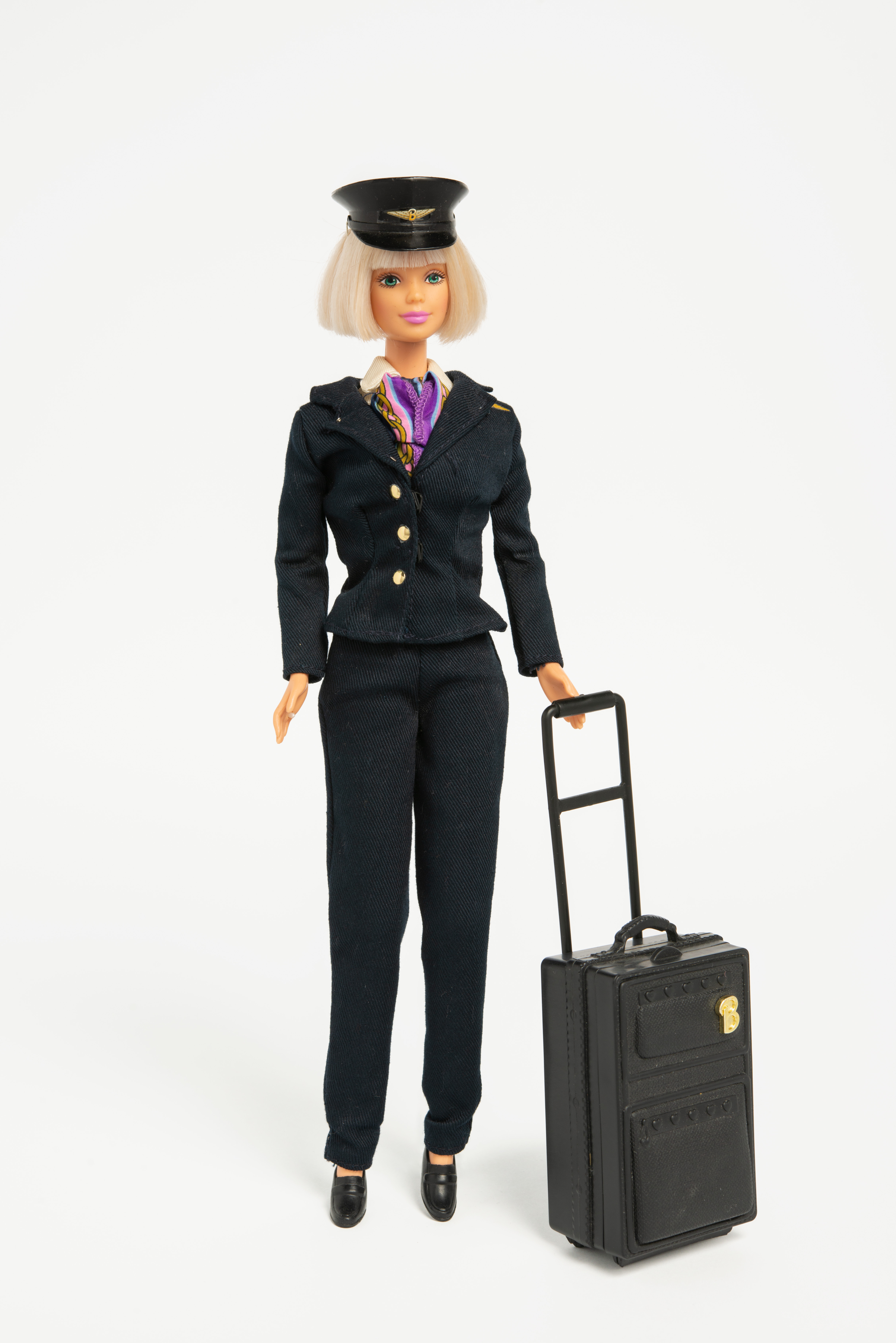 Grafika artykułu: Na zdjęciu lalka Barbie, lalka jest ubrana w strój pilotki, na walizkę na kółkach i czapkę pilota.