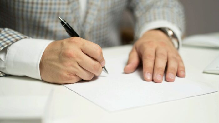 Grafika artykułu: na zdjęciu dłonie starszej osoby podpisujące coś na kartce papieru.
