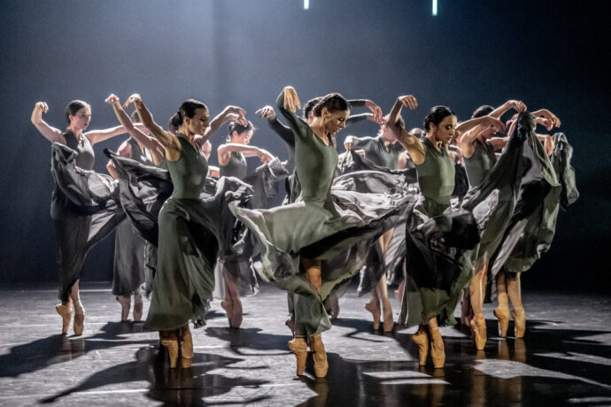 Grafika artykułu: na zdjęciu kilkanaście baletnic, stoją w pozycji baletowej ze skrzyżowanymi nogami i rękami uniesionymi w górę, ubrane w szare, długie suknie, od góry pada na nie jasne, białe światło