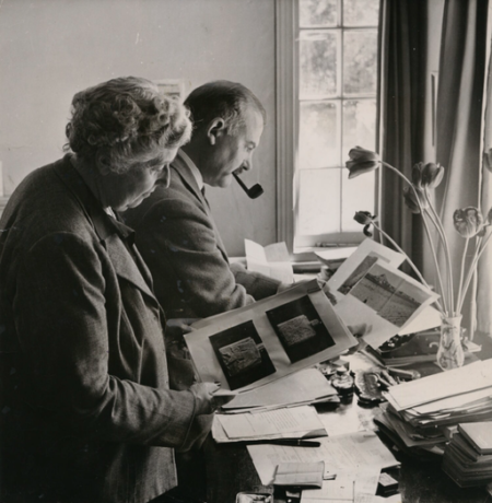 Grafika artykułu: archiwalne, czarno-białe zdjęcie, Agatha Christie i Max Mallowan w 1950 roku, kobieta i mężczyzna  stoją przy biurku, oglądają zdjęcia, mężczyzna ma w buzi fajkę. 