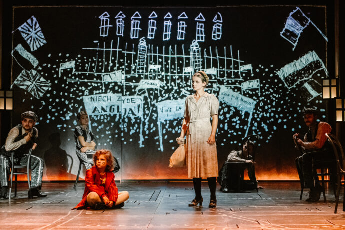 Grafika artykułu: na scenie aktorzy Teatru Muzycznego podczas spektaklu "Irena", w centrum stoi kobieta w białej sukni, trzyma worek, patrzy w lewą stronę, po lewej stronie siedzi dziecko w czerwonym płaszczu, w tle inscenizacja obozowa.