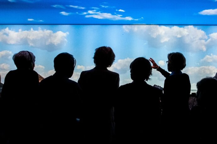Grafika artykułu: na zdjęciu osoby zwiedzające wystawę, znajdują się w cieniu, zwrócone są tyłem, w tle błękitne niebo z kilkoma chmurami.
