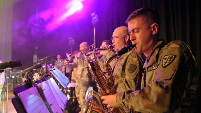 Grafika artykułu: na zdjęciu członkowie zespołu muzycznego NATO, mężczyźni ubrani są w mundury, w dłoniach trzymają różne instrumenty dęte, patrzą na stojące przed nimi stojaki z nutami, oświetla ich fioletowe światło sceniczne.