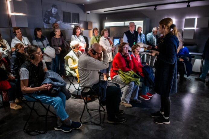 Grafika artykułu: na zdjęciu seniorzy biorący udział w oprowadzaniu po Centrum Szyfrów Enigma, niektórzy siedzą na krzesłach, niektórzy stoją w tyle pomieszczenia, seniorzy zwróceni są w stronę przewodniczki, wszyscy mają zestawy słuchawkowe.