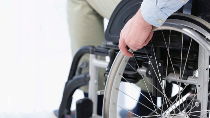 Grafika artykułu: zdjęcie osoby na wózku inwalidzkim — zbliżenie dłoni trzymającej koło wózka inwalidzkiego.
