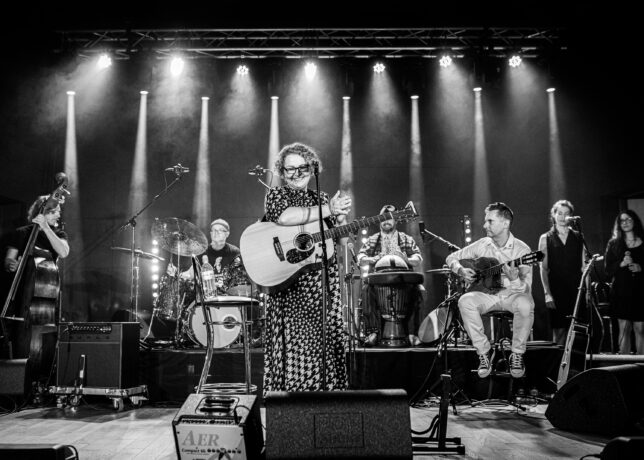 Grafika artykułu: na zdjęciu wokalistka Dorota Józefiak z zespołem na scenie, fotografia jest czarno-biała.