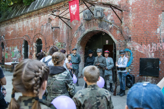 Grafika artykułu: na zdjęciu uczestnicy wydarzeń podczas Dni Twierdzy Poznań, stoją tyłem do obiektywu, przed nimi przewodnicy ubrani w historyczne stroje z czasów wojny, stoją przed wejściem do Fortu II.