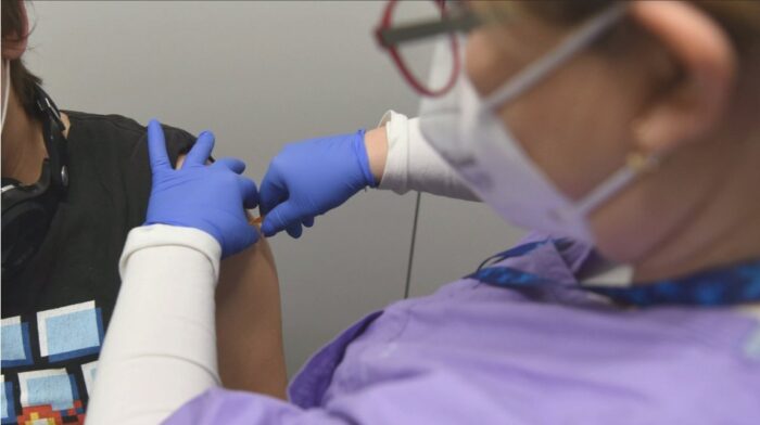 Grafika artykułu: pracowniczka ochrony zdrowia w fioletowym fartuchu i niebieskich rękawiczkach robi zastrzyk w ramię osobie znajdującej się po lewej stronie pola obrazowego.