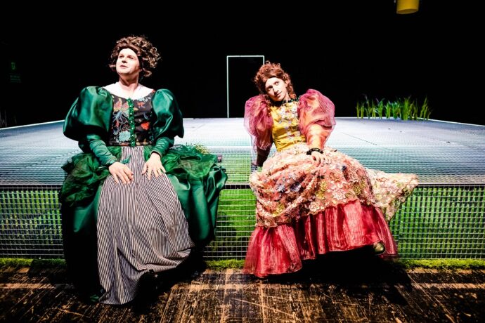 Zdjęcie przedstawia dwóch mężczyzn w kobiecych sukniach, jedna w kolorze zielonym, druga różowa. Mają peruki z długimi włosami. Siedzą na scenie.